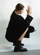 Lade das Bild in den Galerie-Viewer, In einer Bet-Pose hockende Frau, welche einen tiefen Pferdeschwanz, silberfarbenen Schmuck sowie einen schwarzen oversized Mantel trägt. An ihren Füßen befinden sich schwarze, auffällige Leder-Stiefeletten von Cedoublé. Diese Schuhe sind neben der Schnürung mit einem Reißverschluss ausgestattet. Highlights sind die roséfarbenen Steppnähte sowie der prägnante Absatz.