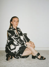 Lade das Bild in den Galerie-Viewer, Eine Frau mit langen Haaren im Sleek-Look trägt ein schwarz-weißes Kleid mit grafischen Mustern und eine elegante Absatzsandale aus weich gepolstertem, schwarzen Leder mit zwei Riemen. Der obere Riemen hat einen Klettverschluss