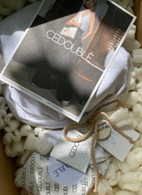 Load image into Gallery viewer, Blick in den Versandkarton von Cedoublé. Zu sehen ist die schöne Verpackung mit einem weißen Baumwollbeutel sowie ein getöpferter Anhänger mit positiver Affirmation. Obenauf liegt eine handgeschriebene Grußkarte des Herstellers. Sonne fällt sanft in das Paket. Das Foto zeigt exemplarisch die liebevolle Verpackung, die Kunden und Kundinnen nach Bestellung erhalten.
