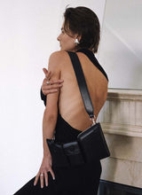 Lade das Bild in den Galerie-Viewer, Foto eines weiblichen Models, welche ein schwarzes Kleid mit einem tiefen Rückenausschnitt trägt und mit dem Rücken zur Kamera gewandt vor einem edlen Kamin steht. Das Model trägt um die linke Schulter eine schwarze, schlanke Ledertasche, welche in drei Fächer unterteilt ist. Die Tasche ist von der Marke Cedoublé.