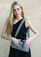 Lade das Bild in den Galerie-Viewer, Bild eines weiblichen Models mit langen, blonden Haaren, gekleidet in einem schwarzen, langen Kleid mit seitlichem Cut-out rechts. Eine graue, genarbte Umhängetasche des Labels Cedoublé hängt ihr frontal über die linke Schulter, mit den Händen deutet die Frau ein Öffnen des mittleren Fachs mit Druckknopf an. Der Blick des Models geht direkt in die Kamera, er ist ernshaft.