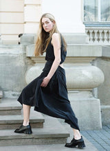 Lade das Bild in den Galerie-Viewer, Bild eines Models mit langen, blonden Haaren, welches mit ernstem Blick die Treppe eines historischen Gebäudes hinauf schreitet. Das schwarze, lange Kleid verfügt über einen Cut-out am Rücken. An den Füßen trägt die Dame extravagante Leder-Schnürschuhe mit einem ausdrucksstarken Absatz. Der Kontrast zwischen femininer Kleidung und klobigeren Schuhen ist hier sehr schön gelungen.