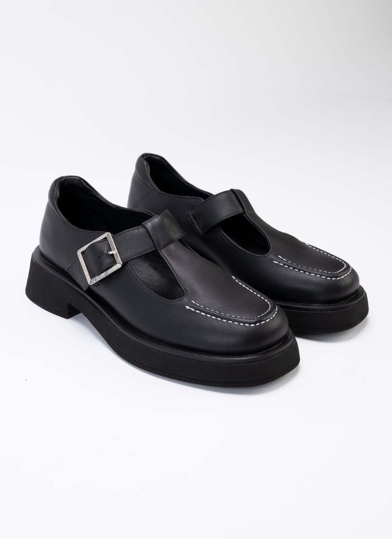 Foto eines Paares chunky Mary Jane Schuhe aus hochwertigem, schwarzem Leder. Die Schuhe sind mit schwarzem Leder gefüttert, die Laufsohle mit dezentem Plateau ist schwarz, die Schnalle eckig und silber. Typisch für diesen Schuhstyle gibt es seitlich auf dem Schaft verlaufende Aussparungen, die den Blick auf einen Teil des Fußes freilegen.