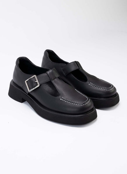 Foto eines Paares chunky Mary Jane Schuhe aus hochwertigem, schwarzem Leder. Die Schuhe sind mit schwarzem Leder gefüttert, die Laufsohle mit dezentem Plateau ist schwarz, die Schnalle eckig und silber. Typisch für diesen Schuhstyle gibt es seitlich auf dem Schaft verlaufende Aussparungen, die den Blick auf einen Teil des Fußes freilegen.