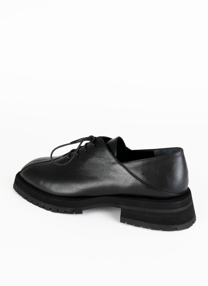Produktfoto eines burschikosen Unisex-Schnürschuhs aus Leder mit Oxford-Schnürung und ausgeprägter Laufsohle in schwarz. Eine asymmetrische Naht ziert die Schuhfront