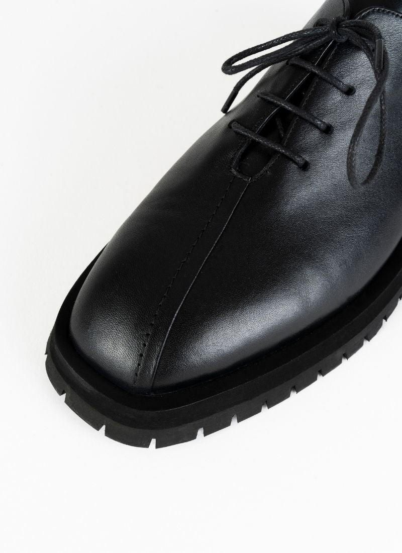 Detailfoto eines burschikosen Leder Derbys mit Oxford-Schnürung und ausgeprägter Laufsohle in schwarz. Eine asymmetrische Naht ziert die Schuhfront