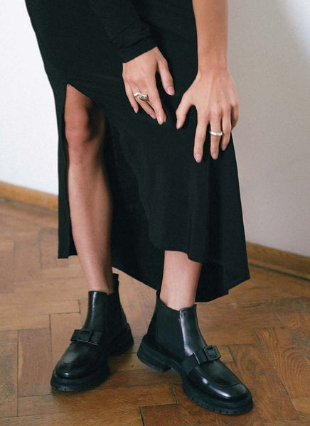 Ein Foto von grazilen Modelbeinen, die in schwarzen Leder Chelsea Boots mit Schleife stecken. Das Model ist sehr edel mit einem langen, schwarzen Kleid mit Schlitz gekleidet. Die Frau zieht mit gepflegten Händen, die von silbernen Ringen geziert werden, das Kleid leicht nach oben, sodass der Fokus auf die hochwertigen Schuhe gelenkt wird. Das Model befindet sich in einem Altbau, es ist ein schönes Fischgrät-Parkett zu sehen.