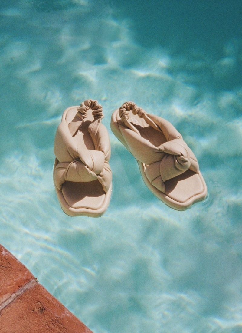 Foto von einem Paar cremefarbener Ledersandalen, das auf einer Pool-Oberfläche schwimmt. Diese Sandalen verfügen über ein modisches Knotendetail und sind voluminös. An der Ferse befindet sich ein mit Leder ummantelter Fersen-Strap, der elastisch ist. Die Schuhspitze ist karreeförmig und besticht durch eine kleine Einkerbung. Die Schuhe sind von Cedoublé und erinnern an ein Modell von MIU MIU by Prada.