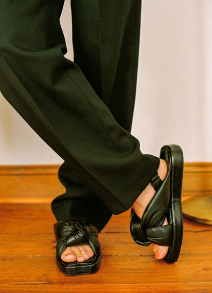 Überkreuzte Frauenbeine mit lässiger Hose. An den Füßen sind schwarze chunky Ledersandalen mit großem verschlungenem Knoten.