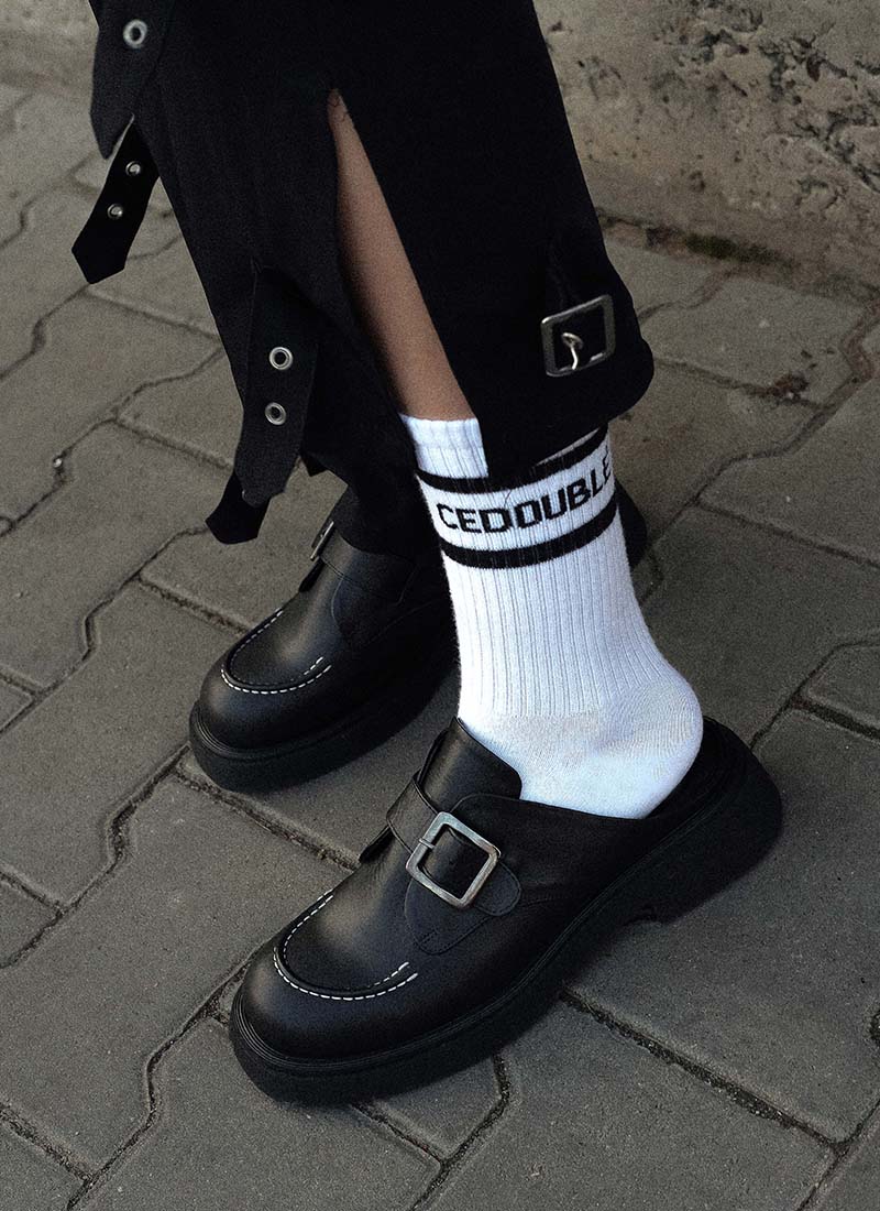 Bild einer coolen Kombination von weißen Tennissocken mit Logo-Print und hochwertigen schwarzen Leder-Clogs mit weißen Nähten. Die schwarze Hose, die das Model dazu trägt, verfügt über einen Beinschlitz. Dieser klafft offen, sodass der Logo-Schriftzug auf der Socke gut zur Geltung kommt. Sowohl Socke als auch Schuhe sind von der Marke Cedoublé.  Die Clogs verfügen über eine Monk-Schnalle und stehen optische großen Designerlabels in nichts nach.