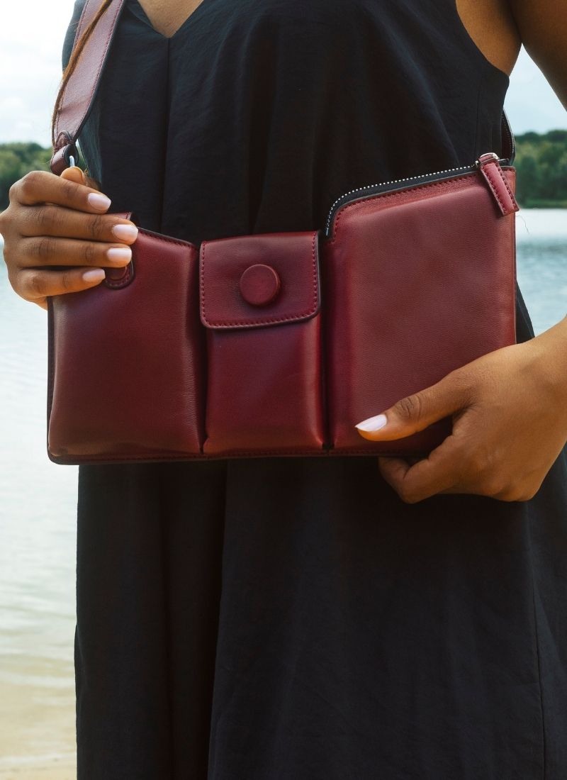 Das Foto zeigt den Oberkörper eines Models mit einem weiten, schwarzen Kleid. Zu sehen sind zwei gepflegte Hände, die eine rote Crossbody Bag mit drei Abteilen umfassen. Die rote Tasche hängt locker am Körper und schmiegt sich den Körperkonturen an. Sie ist von der Marke Cedoublé und wurde aus hochwertigem Lammleder gefertigt.