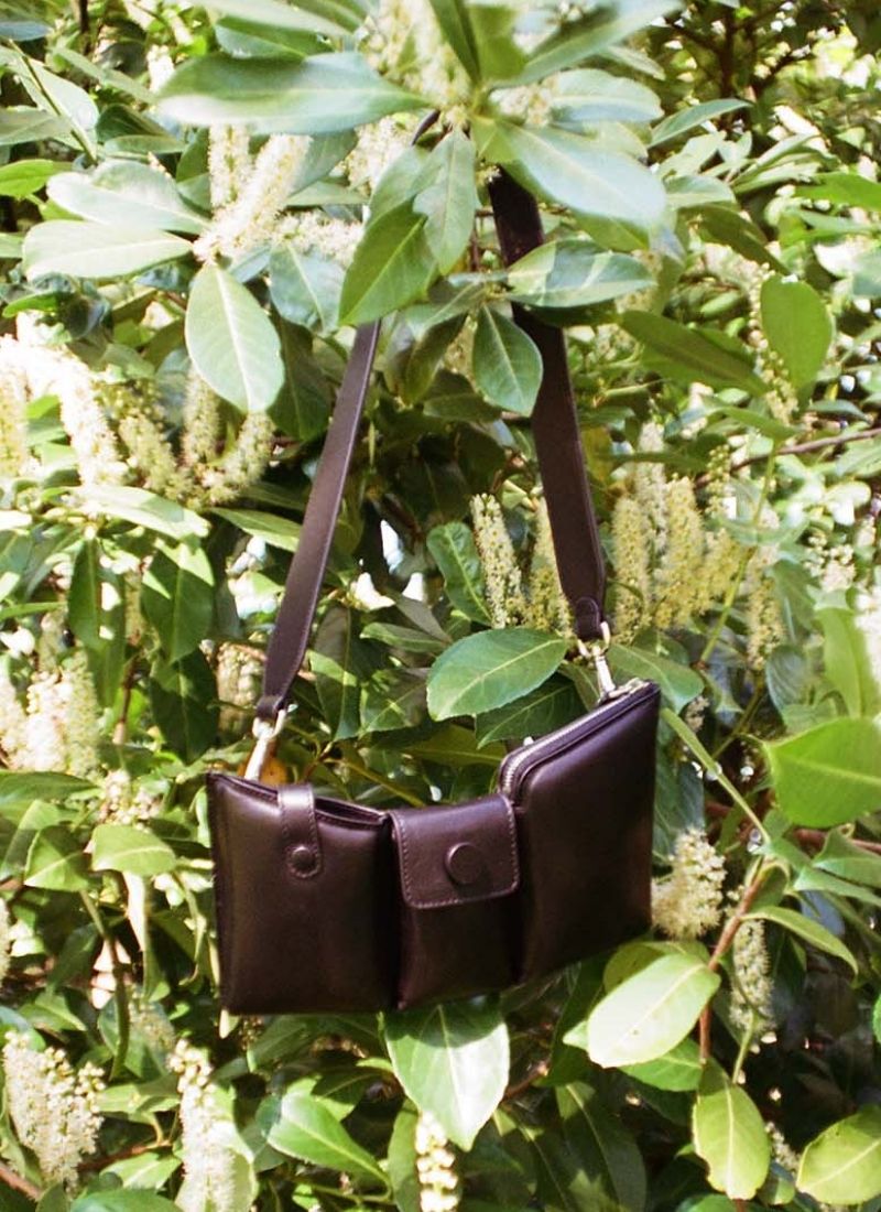 Puffy black leather bag by CEDOUBLÉ hanging in a bush with white blossoms on a sunny day 