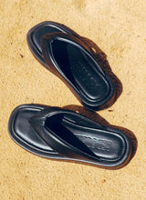 Load image into Gallery viewer, In der prallen Sonne stehen ein Paar Zehensteg-Sandalen aus schwarzem Glattleder auf einem sandigen Untergrund. Die Schuhe sehen aus wie Designer Sandalen. Die weich gepolsterten Lederriemen im V-Schnitt, der Zehensteg sowie das große Hersteller-Logo von Cedoublé erzeugen einen sehr hochwertigen, coolen Look.
