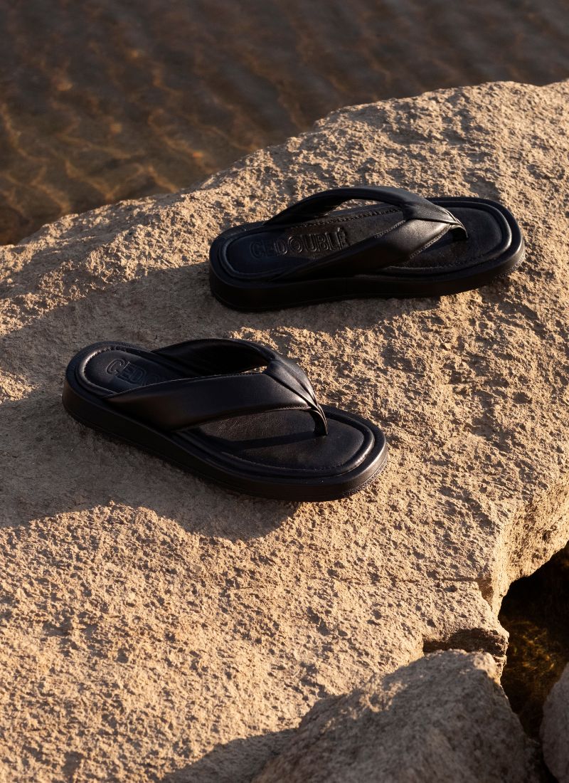 Foto von einem Paar chunky Leder Flipflops in schwarz in einer Urlaubsatmosphäre. Die Sandalen aus Echtleder stehen auf einem sandfarbenem Felsen, im Hintergrund ist Wasser zu sehen. Die Schuhe verfügen über einen Zehensteg und ein großes Logo des Herstellers Cedoublé auf der Decksohle.