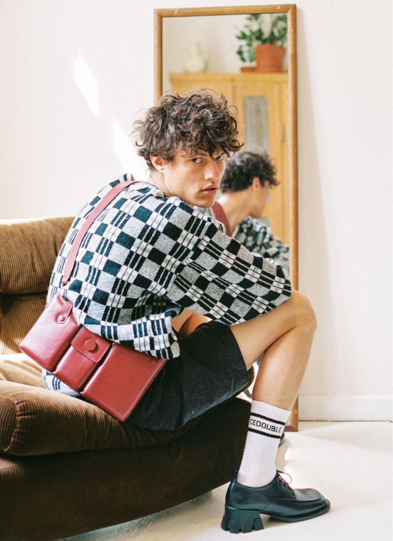 Cooles Foto eines männlichen Models mit lockigen Haaren und modischem Styling auf einem braunen Cordsofa sitzend. Im Fokus stehen die rote Umhängetasche aus feinem Kalbsleder sowie die schwarzen Lederschuhe mit einem ausgefallenen Absatz. Beide Artikel sind vom deutschen Hersteller Cedoublé.